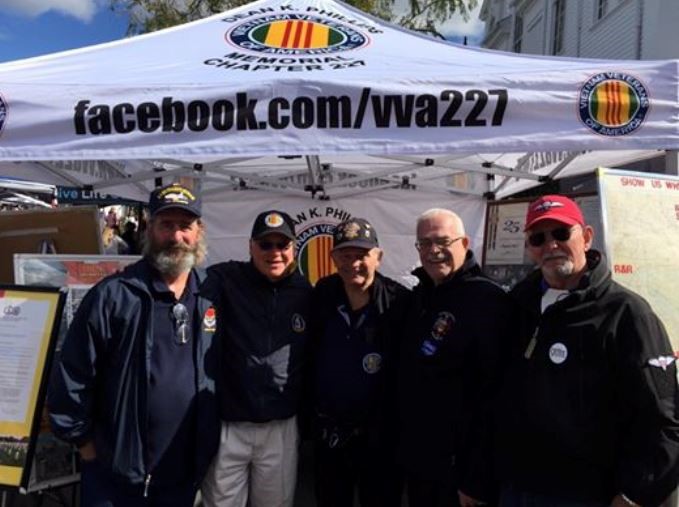Five Chapter 227 members in chapter jackets and caps standing together in front of the chapter's canopy at a community fall festival.