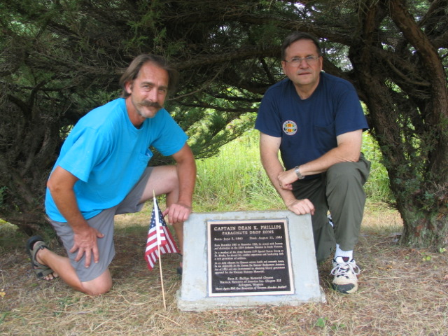Two men kneeling on either side of the newly installed Dean K. Phillips plaque beneath a tree at the drop zone, with a small American flag planted in the grass in front of it.
